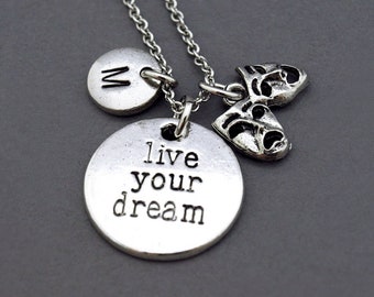 May include: Silver necklace with three charms: a round charm with the letter "W" engraved, a round charm with the text "live your dream", and a charm of two theatrical masks.