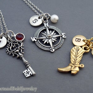 May include: Three silver necklaces with charms. The first necklace has a key charm with a red gemstone, a silver disc with the letter "J" and a silver compass charm. The second necklace has a silver compass charm with a pearl. The third necklace has a gold boot charm, a gold disc with the letter "B" and a blue gemstone.