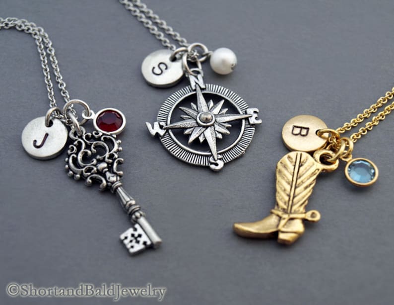 May include: Three silver necklaces with charms. The first necklace has a key charm with a red gemstone, a silver disc with the letter "J" and a silver compass charm. The second necklace has a silver compass charm with a pearl. The third necklace has a gold boot charm, a gold disc with the letter "B" and a blue gemstone.