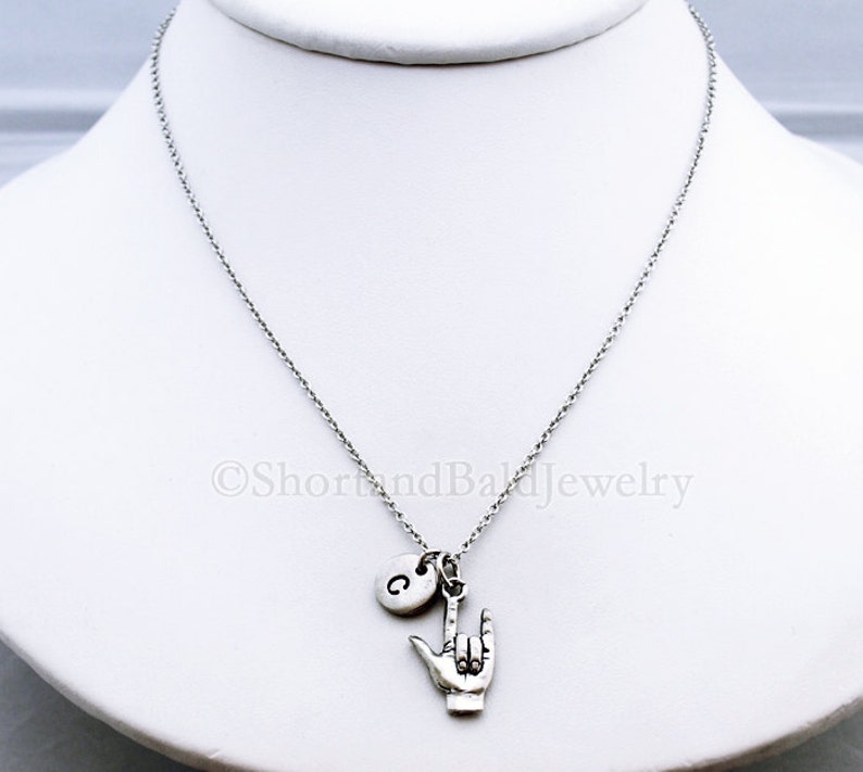 May include: Silver necklace with a charm of a hand making the sign for 'I love you' in American Sign Language. The necklace also has a small charm with the letter 'C' on it.