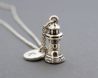 May include: Silver necklace featuring a lighthouse charm and a small disc charm with the letter K. The necklace is displayed against a grey background.