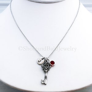 May include: A silver necklace with a silver key charm, a silver initial charm, and a red gemstone charm. The necklace is on a white background.