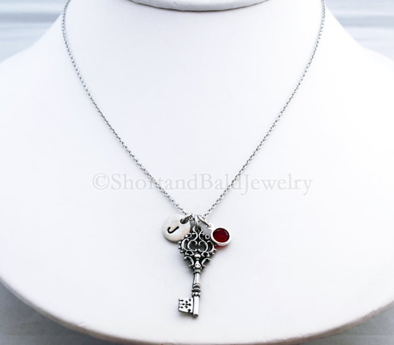 May include: A silver necklace with a silver key charm, a silver initial charm, and a red gemstone charm. The necklace is on a white background.