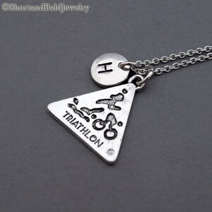 May include: Silver necklace with a triangle pendant featuring a triathlon symbol and the word "TRIATHLON" inscribed on it. The necklace also has a small round charm with the letter "H" on it.