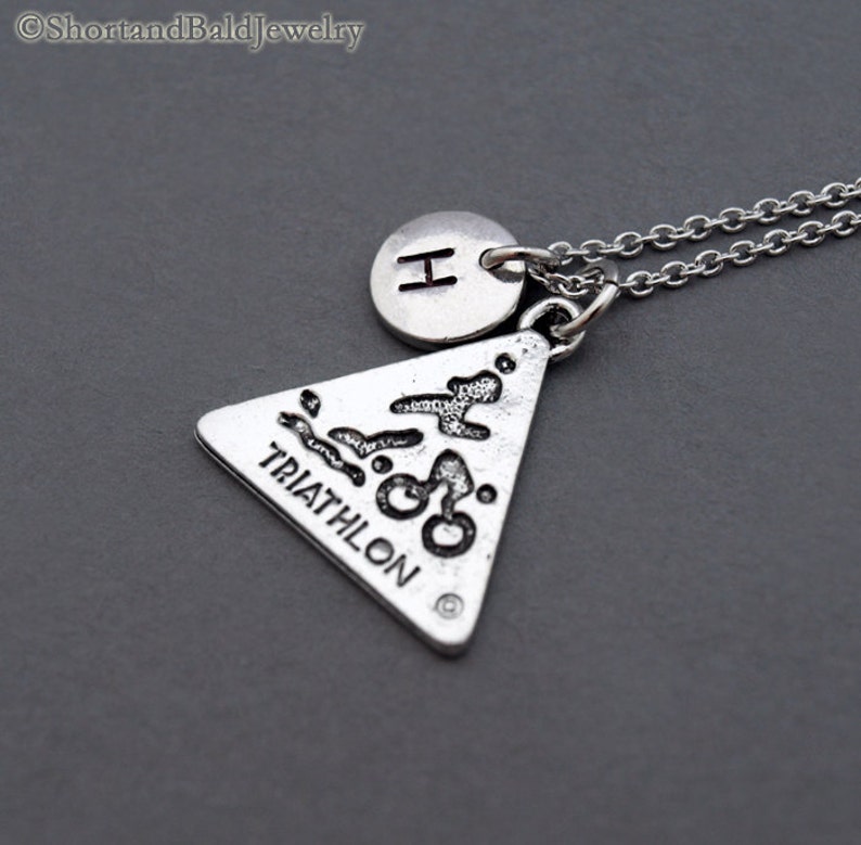 May include: Silver necklace with a triangle pendant featuring a triathlon symbol and the word "TRIATHLON" inscribed on it. The necklace also has a small round charm with the letter "H" on it.