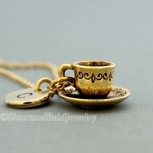 May include: Gold necklace featuring a charm of a miniature teacup and saucer. The teacup has an ornate design around the rim. The necklace also includes a small round charm with the letter C.