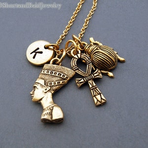 May include: Gold necklace with multiple charms, including a scarab beetle, an ankh, a bust of Nefertiti, and a round charm with the letter "K" on it.