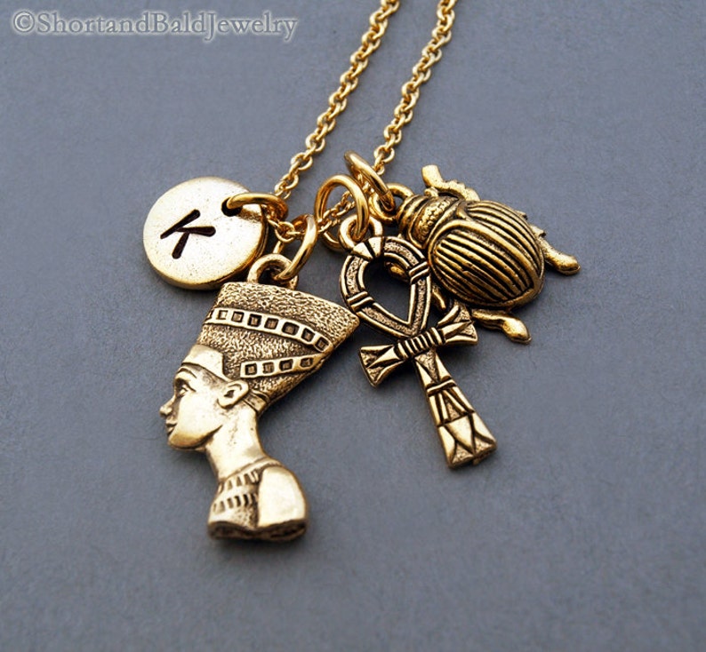 May include: Gold necklace with multiple charms, including a scarab beetle, an ankh, a bust of Nefertiti, and a round charm with the letter "K" on it.