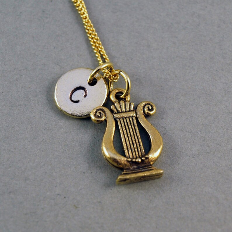 K&ouml;nnte beinhalten: Goldkette mit einem kleinen runden Anh&auml;nger, der mit dem Buchstaben "C" graviert ist, und einem Lyra-Anh&auml;nger.