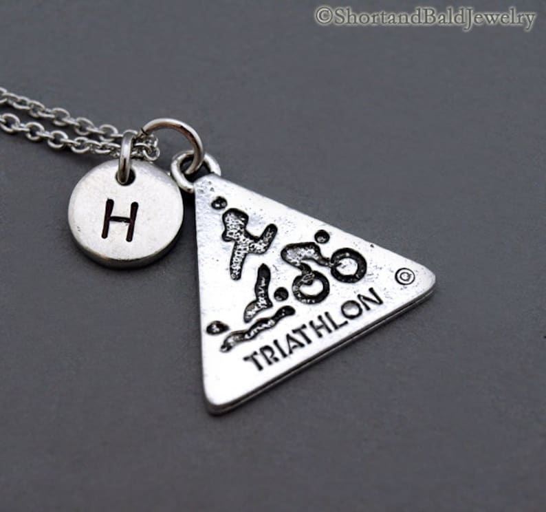 May include: Silver necklace with a triangle pendant featuring a stylized triathlon symbol and the word "TRIATHLON". The necklace also has a small round charm with the letter "H" on it.