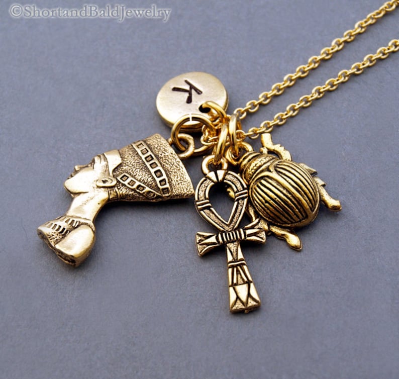 May include: Gold necklace with multiple charms, including an Egyptian queen profile, an ankh, a scarab beetle, and a round charm with the letter "K" on it.