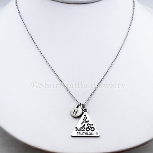 May include: Silver necklace with a triangle pendant featuring a triathlon symbol and a small initial charm. The pendant reads "TRIATHLON".