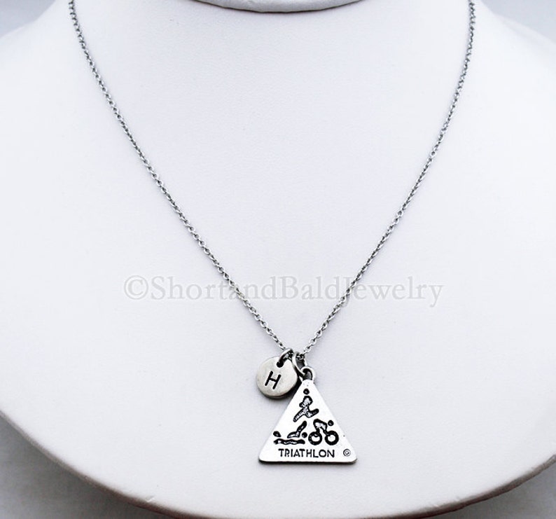May include: Silver necklace with a triangle pendant featuring a triathlon symbol and a small initial charm. The pendant reads "TRIATHLON".
