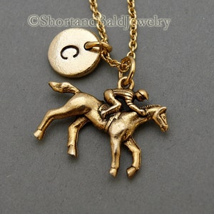 May include: Gold necklace featuring a jockey riding a horse charm and a round initial charm engraved with the letter "C". The necklace is displayed against a grey background.