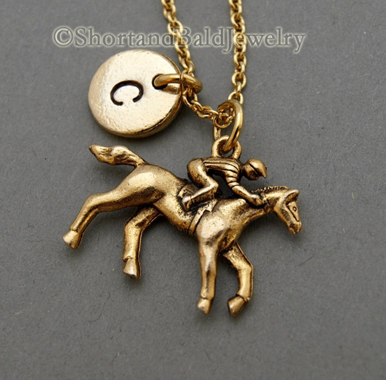 May include: Gold necklace featuring a jockey riding a horse charm and a round initial charm engraved with the letter "C". The necklace is displayed against a grey background.