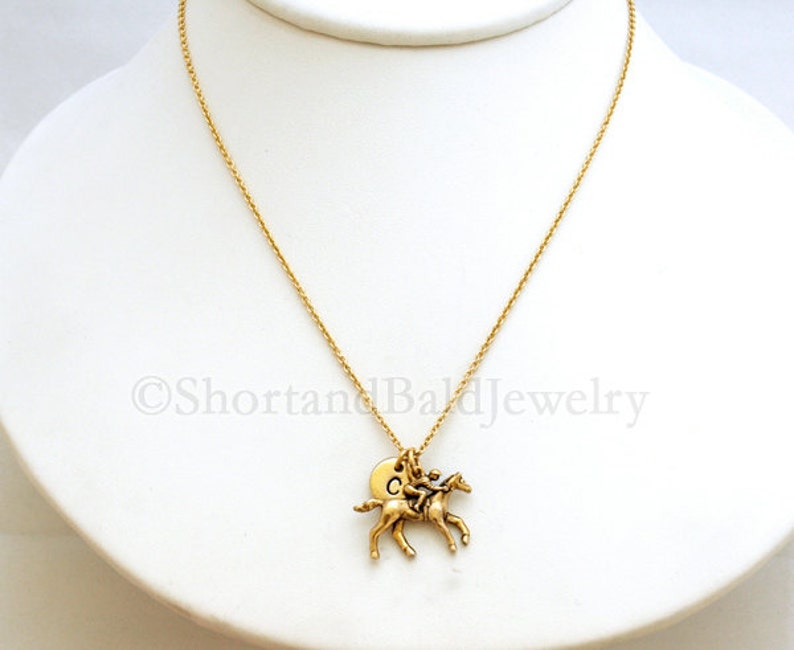 May include: A gold chain necklace with a gold horse and rider charm. The charm has a small round disc with the letter "C" on it.