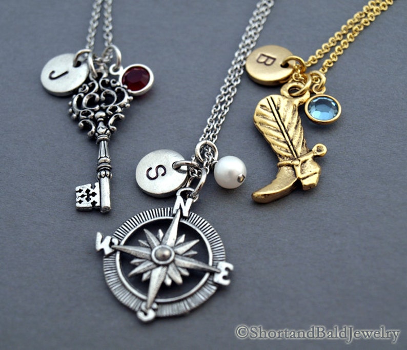May include: Three silver necklaces with charms. The first necklace has a silver key charm, a silver initial charm with the letter "J", and a red gemstone. The second necklace has a silver compass charm, a silver initial charm with the letter "S", and a white pearl. The third necklace has a gold boot charm, a gold initial charm with the letter "B", and a blue gemstone.