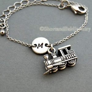May include: Silver charm bracelet with a train charm and a round charm with the letter "M" engraved on it.  ShortandBaldjewelry is printed on the background.
