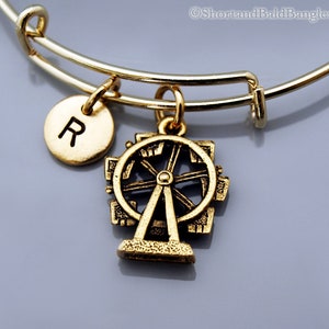 May include: Gold-toned expandable bangle bracelet with a Ferris wheel charm and a round charm with the letter "R". The Ferris wheel charm is detailed with spokes and cabins. The bracelet is on a gray background.
