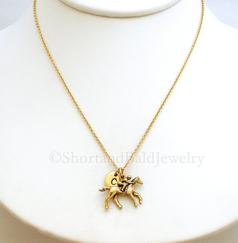 May include: Gold necklace featuring a jockey riding a horse charm and a personalised initial disc charm. The initial disc charm is engraved with the letter "C".