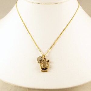 K&ouml;nnte beinhalten: Eine goldene Kettenkette mit einem kleinen goldenen Leier-Anh&auml;nger und einem goldenen Initial-Scheibenanh&auml;nger.