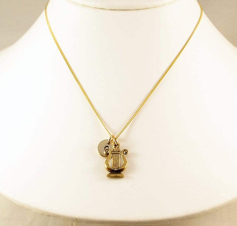 K&ouml;nnte beinhalten: Eine goldene Kettenkette mit einem kleinen goldenen Leier-Anh&auml;nger und einem goldenen Initial-Scheibenanh&auml;nger.