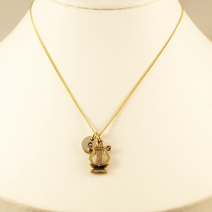 K&ouml;nnte beinhalten: Eine goldene Kettenkette mit einem goldenen Leier-Anh&auml;nger und einem goldenen Initial-Anh&auml;nger. Die Kette befindet sich auf einer wei&szlig;en Puppe.