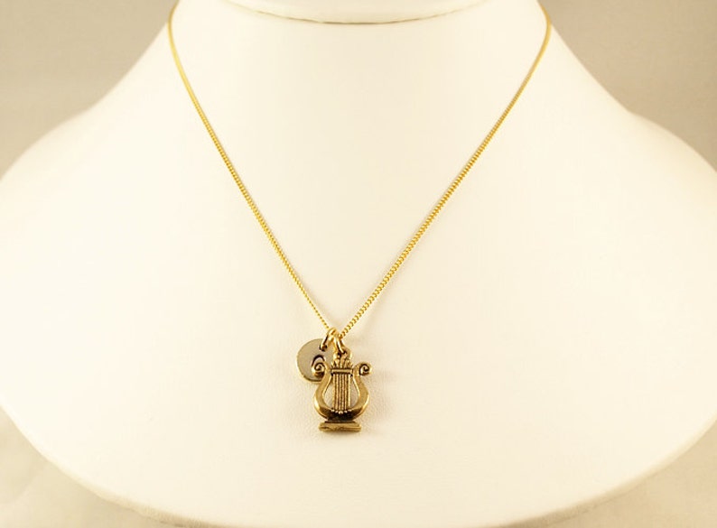K&ouml;nnte beinhalten: Eine goldene Kettenkette mit einem goldenen Leier-Anh&auml;nger und einem goldenen Initial-Anh&auml;nger. Die Kette befindet sich auf einer wei&szlig;en Puppe.