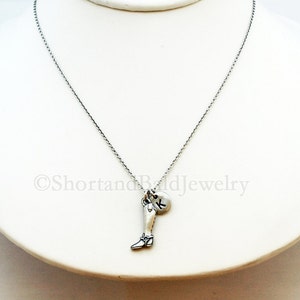 May include: A silver chain necklace with a charm of a leg wearing a boot and a silver disc with the letter K.