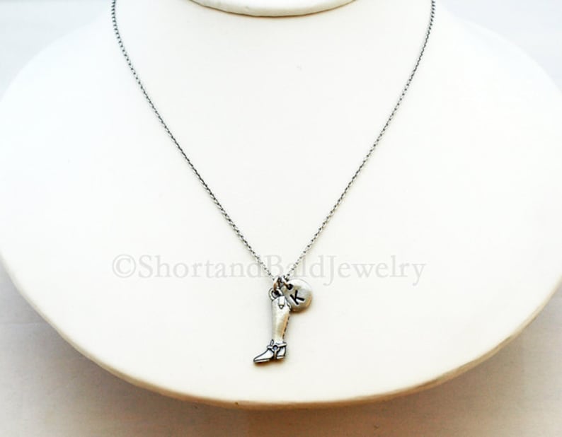 May include: A silver chain necklace with a charm of a leg wearing a boot and a silver disc with the letter K.