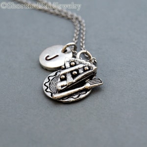 May include: Silver necklace with a pie charm and a letter J charm. The pie charm is a slice of pie on a plate with a fork.