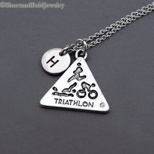 May include: Silver necklace with a triangle pendant featuring a triathlon symbol and the word "TRIATHLON". A small silver circle with the letter "H" is attached to the chain.