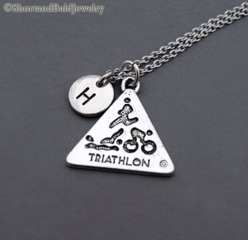 May include: Silver necklace with a triangle pendant featuring a triathlon symbol and the word "TRIATHLON". A small silver circle with the letter "H" is attached to the chain.