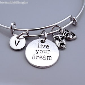 Könnte beinhalten: Silbernes Armband mit einem Anhänger, auf dem "live your dream" steht, und einem Anhänger mit zwei Theatermasken. Das Armband hat außerdem einen Anhänger mit dem Buchstaben "V".