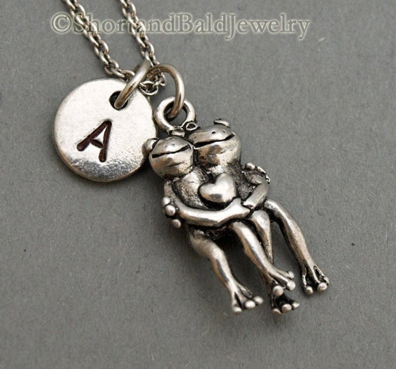 May include: Silver necklace with a charm of two frogs holding a heart. The necklace also has a silver disc charm with the letter A engraved on it.