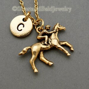May include: Gold necklace featuring a jockey riding a horse charm and a gold initial charm with the letter C. The necklace is displayed against a grey background.