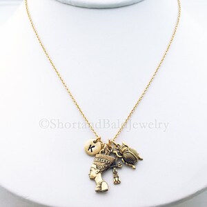 May include: Gold chain necklace with a charm of an Egyptian queen, a scarab beetle, a cross, and a round charm with the letter K.