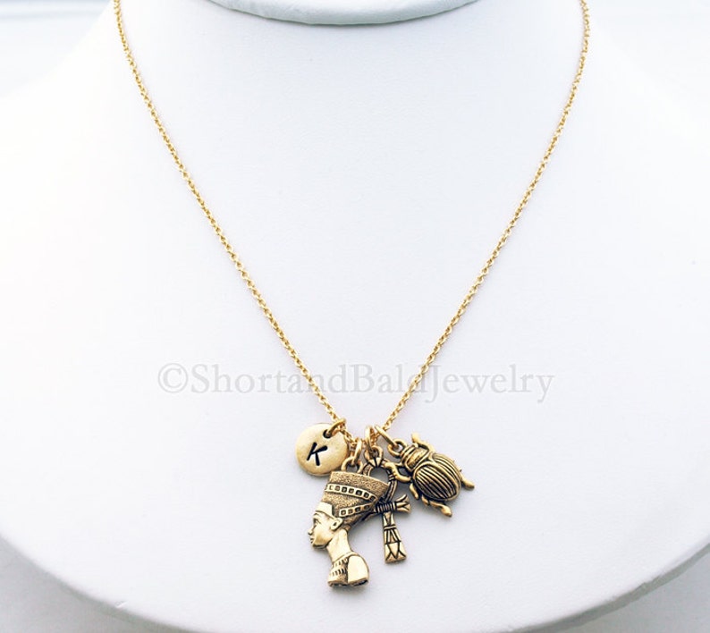 May include: Gold chain necklace with a charm of an Egyptian queen, a scarab beetle, a cross, and a round charm with the letter K.