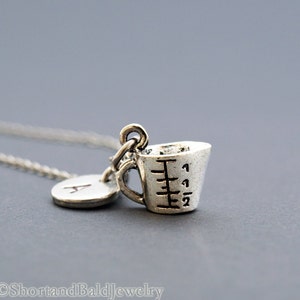 May include: Silver necklace with a charm of a measuring cup and a letter A charm. The measuring cup charm has markings for 1/2, 1, 1 1/2, and 2.