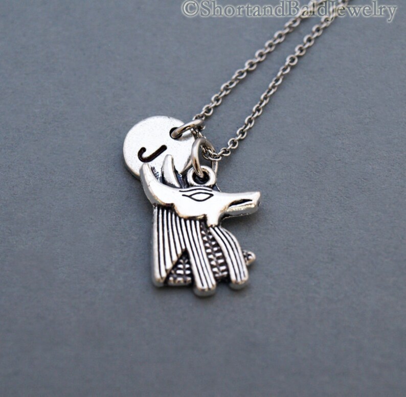 May include: Silver necklace with a charm of an Anubis head and a round charm with the letter J.