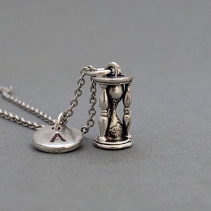 May include: A silver necklace with a small hourglass charm and a round charm with the letter 'A' on it.
