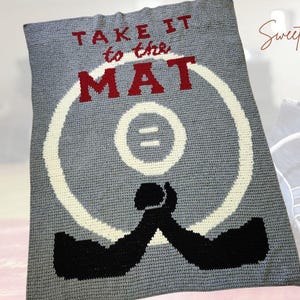 Könnte beinhalten: Eine graue Häkeldecke mit den Worten "TAKE IT to the MAT" in roten Buchstaben. Die Decke zeigt ein Wrestling-Matten-Design mit schwarzen Armen, die sich umarmen. Ein Wrestlingschuh ist im Hintergrund zu sehen.