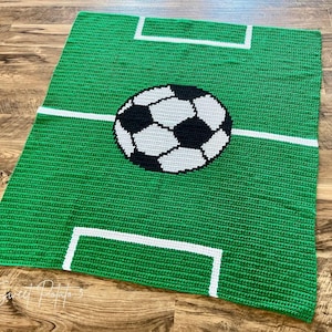 Puede incluir: Una manta verde tejida a ganchillo con un diseño de campo de fútbol. La manta tiene un fondo verde con líneas blancas que marcan los límites del campo y un gran balón de fútbol en el centro. La manta es rectangular.