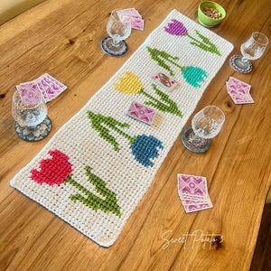 May include: A white crochet table runner featuring colorful tulip designs in red, yellow, blue, teal, and purple. The runner is placed on a wooden table, with playing cards, glasses, and a bowl of nuts.