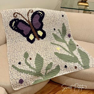 May include: A crocheted blanket with a butterfly design in purple, brown, and yellow. The blanket also has green leaf and floral accents. The blanket is beige and is draped over a beige sofa.