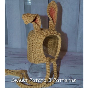 May include: A crocheted bunny hat with long ties. The hat is tan with pink ears. The hat is sitting on a glass surface. "Sweet Potato 3 Patterns" is written on the surface below the hat.