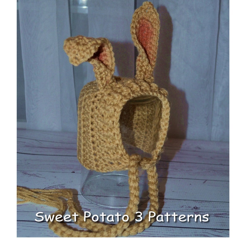 May include: A crocheted bunny hat with long ties. The hat is tan with pink ears. The hat is sitting on a glass surface. "Sweet Potato 3 Patterns" is written on the surface below the hat.