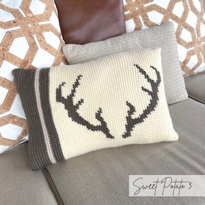 May include: A white crocheted pillow with a brown deer antler design. The pillow has a brown and white striped border. The pillow is sitting on a brown couch.