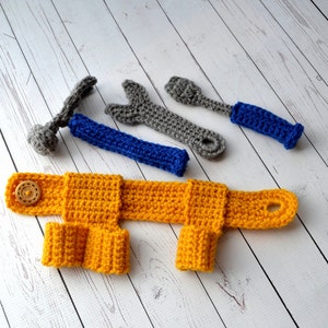 May include: A set of crocheted toy tools, including a hammer, wrench, and screwdriver, in shades of gray and blue. The tools are laid out on a white wooden surface. A yellow crocheted toy vest with a button is also included.