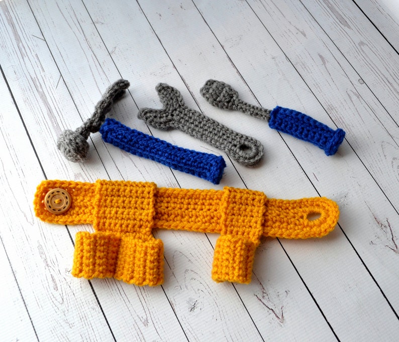 May include: A set of crocheted toy tools, including a hammer, wrench, and screwdriver, in shades of gray and blue. The tools are laid out on a white wooden surface. A yellow crocheted toy vest with a button is also included.